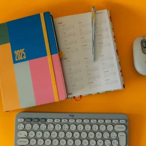 Photo de fournitures de bureau vendues à la Papeterie Valles à Lourdes (65100) dans les Hautes-Pyrénées. Un agenda papier, un stylo, un clavier et une souris d'ordinateur posés sur un fond orange.